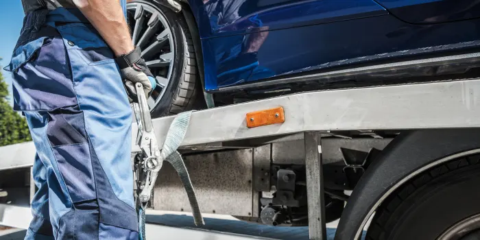 A tow truck worker in blue jeans is strapping a blue car to a flat bed tow truck